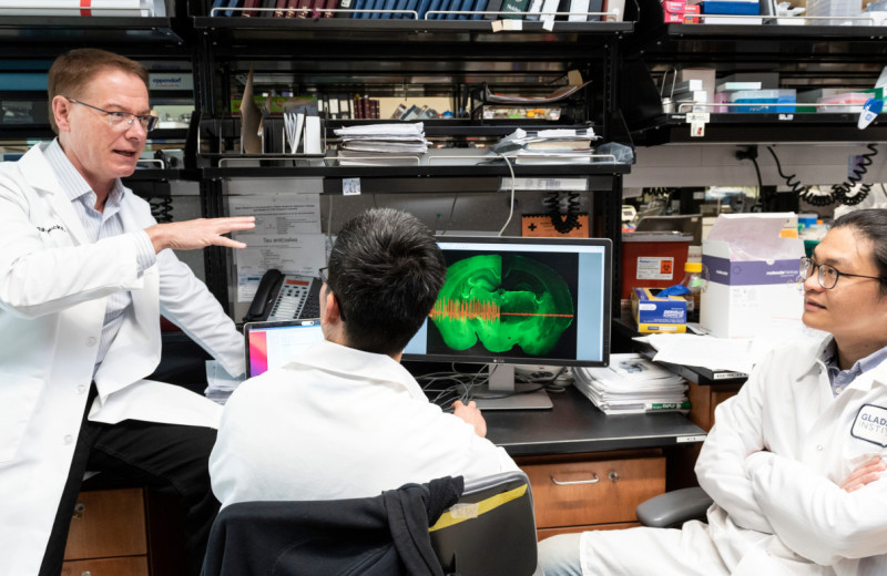 Lennart Mucke with a lab coat, sitting on a lab bench talking to two other scientists who are also seated. 