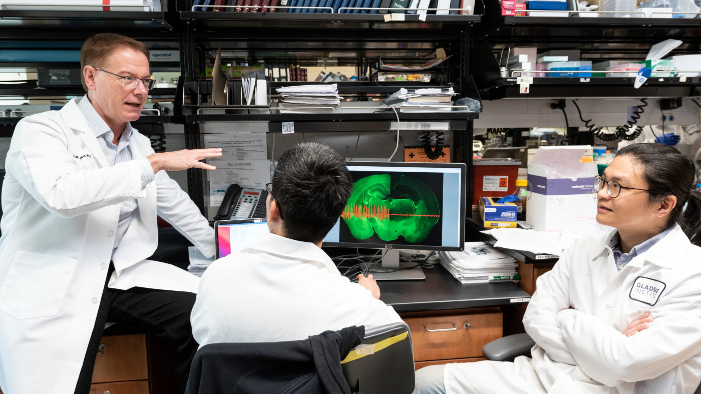 Lennart Mucke with a lab coat, sitting on a lab bench talking to two other scientists who are also seated. 