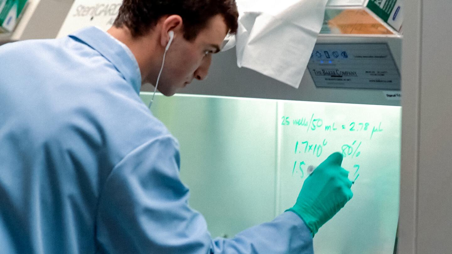 A researcher in a blue lab coat and teal gloves writes calculations on the glass panel of a biosafety cabinet in a laboratory settin