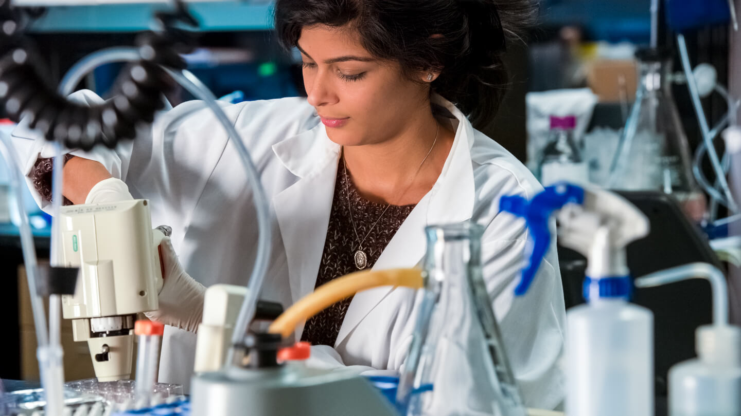 A researcher in a white lab coat and gloves works intently with laboratory equipment, surrounded by glassware, tubing, and supplies in a busy research lab.