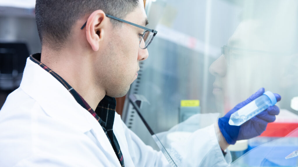 A researcher in a white lab coat and glasses examines a sample tube inside a biosafety cabinet.