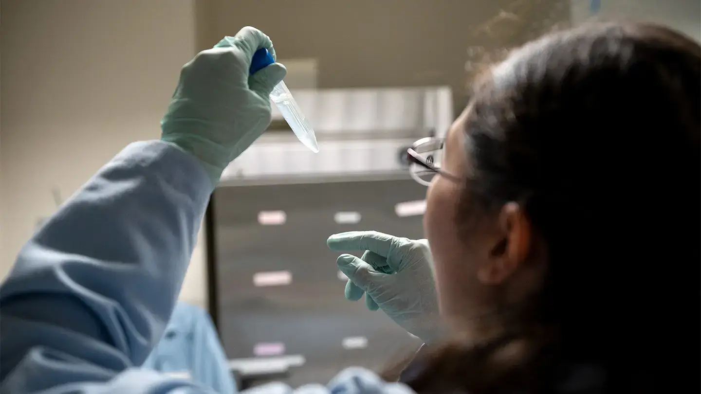 A researcher in a blue lab coat and teal gloves examines a small clear sample vial held up to the light in a laboratory setting.