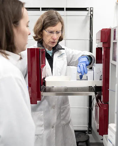 Two scientists in lab coats and gloves loading a piece of lab equipment.