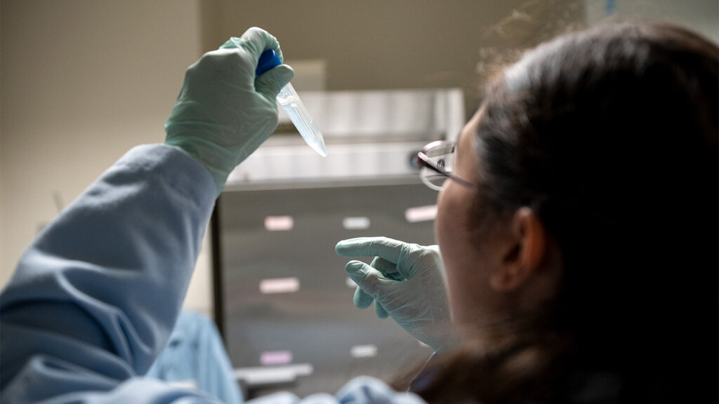 A researcher in a blue lab coat and teal gloves examines a small clear sample vial held up to the light in a laboratory setting.