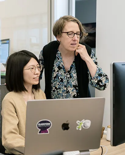 Katie Pollard standing, looking at a computer screen at a desk with another scientist seated in front of the computer