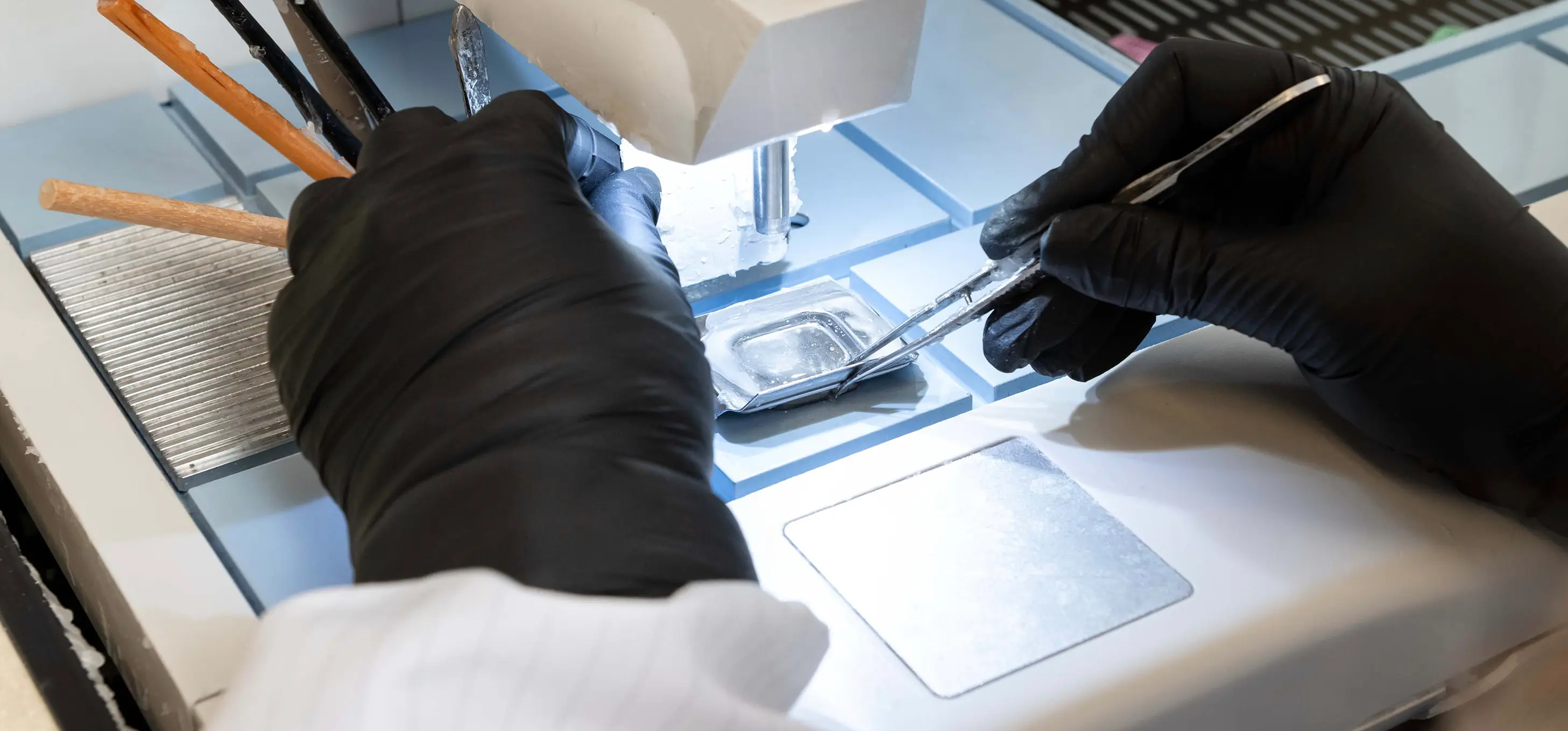 A researcher in black gloves uses tweezers to handle a sample under a microscope in a laboratory setting.