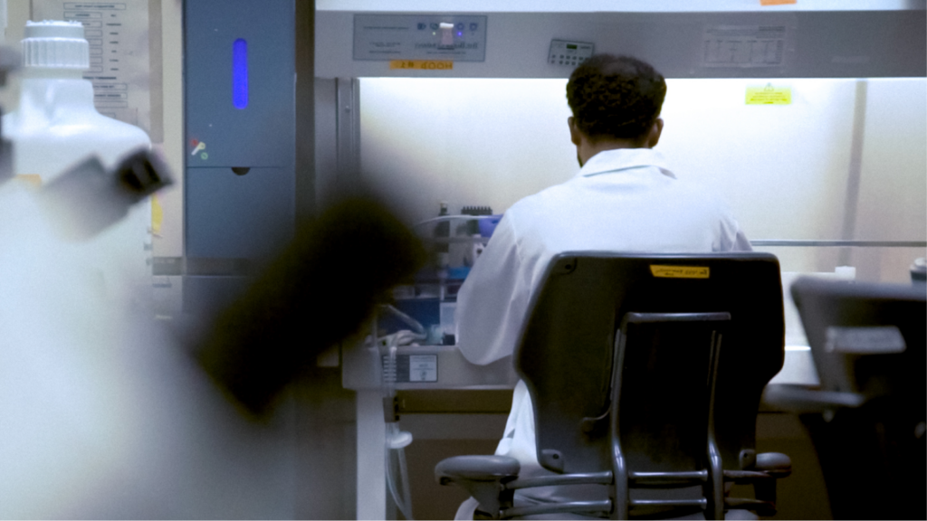 A researcher in a lab coat works at a biosafety cabinet, surrounded by scientific equipment and materials.
