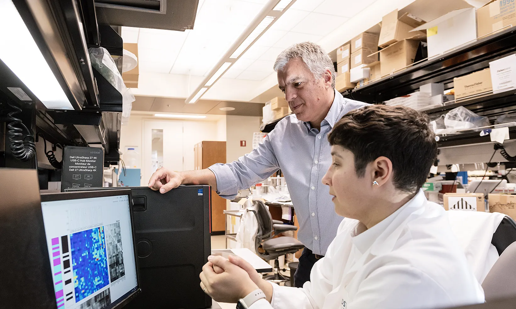 Jorge Palop standing at a lab bench looking at a computer screen with another scientist sitting in front of the computer