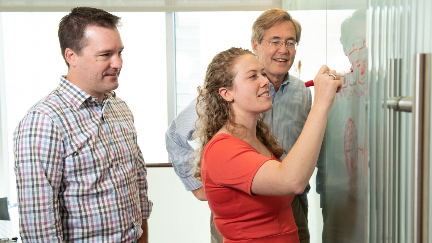 Gladstone Scientist Ashley Libby writing on a white board with Todd McDevitt and Bruce Conklin watching