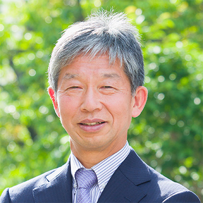 A smiling man in a suit stands outdoors, surrounded by greenery, representing the 2026 Ogawa-Yamanaka Stem Cell Prize Reception.